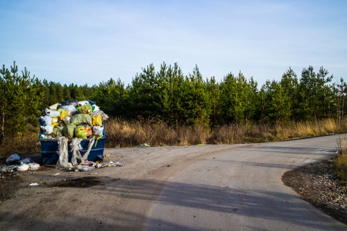 An overflowing blue skip positioned on the edge of a dirt road in a rural area, filled with a variety of waste materials including plastic bags, cardboard boxes, and miscellaneous rubbish, with some debris scattered on the ground in front of it. Behind the skip, there is a line of green trees and shrubbery under a clear blue sky, indicating an outdoor environment near Kensington. The scene suggests rubbish accumulation requiring professional clearance services, such as those provided by Garden Clearance Kensington, within the local context of the postcode area associated with Kensington or nearby London districts.