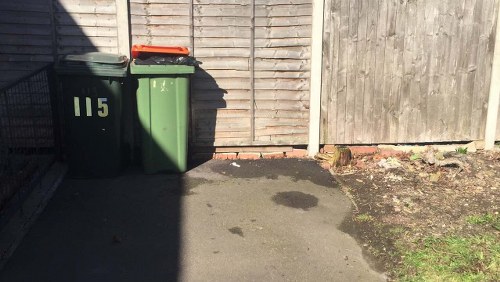 The image shows two large wheelie bins positioned on a paved driveway adjacent to a wooden fence, with the left bin being green and marked with the number 115, and the right bin in a darker shade of green. The bins are placed close together, with the green bin slightly in front of the other, and both are intended for rubbish or waste collection. There is a dark shadow cast across the lower left part of the driveway, likely caused by a nearby structure or fence, creating a stark contrast with the sunlit area. The pavement appears to have some dark patches or stains, and the fence behind the bins is made of vertical wooden planks, showing weathering and natural wood tones. To the right, part of the garden or external area is visible, with dirt, small patches of grass, and minimal vegetation, typical of a residential setting in the vicinity of Kensington. This scene exemplifies typical rubbish disposal arrangements in a UK suburban or residential property, as handled by local rubbish removal services such as Garden Clearance Kensington.