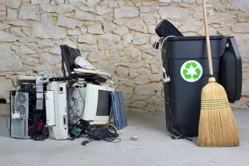 A pile of discarded electronic waste, including computer towers, monitors, and various cables, stacked irregularly on a paved surface against a stone wall. To the right, there is a large grey rubbish bin with a green recycling symbol sticker, a black open lid, and a wooden broom leaning against it, indicating a waste collection or rubbish clearance setting in an outdoor area. The scene reflects typical waste management items that Garden Clearance Kensington might handle in a residential or commercial garden clearance or rubbish removal service within Kensington or nearby London neighborhoods.