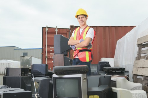 A construction worker wearing a yellow safety helmet and a high-visibility vest stands outdoors on a yard or storage area, surrounded by discarded electronic equipment such as old computer monitors, keyboards, and other obsolete electronic devices. In the background, there is a large shipping container and industrial buildings, indicating a waste collection or disposal site commonly used for rubbish removal services in the Kensington area. The worker is smiling and has his arms crossed, with the electronic waste placed in front of him on the ground, illustrating a situation of electronic waste clearance or recycling. The environment features a cloudy sky and a clean, organized space dedicated to waste management activities. This scene reflects professional rubbish removal operations, specifically focusing on the collection and disposal of electronic waste as part of a comprehensive rubbish clearance service in Kensington by Garden Clearance Kensington.