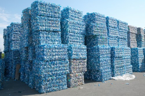 A large outdoor storage area containing numerous stacked pallets of compressed plastic bottles, predominantly blue and clear, with some white labels visible. The bottles are tightly packed together in neat, rectangular formations, creating a substantial wall of recyclable materials. The stacks are arranged in rows on a paved surface, with some pallets leaning slightly. In the background, a pale blue sky is visible, and a few scattered bottles and small pieces of plastic are lying on the ground near the stacks. This scene reflects typical waste management practices related to plastic recycling, relevant to rubbish removal services such as those offered by Garden Clearance Kensington. The image emphasizes the scale and organization of bulk plastic waste, situated in an environment that could be part of an industrial or waste disposal facility in or near London, within the postcode area covered by Kensington services.
